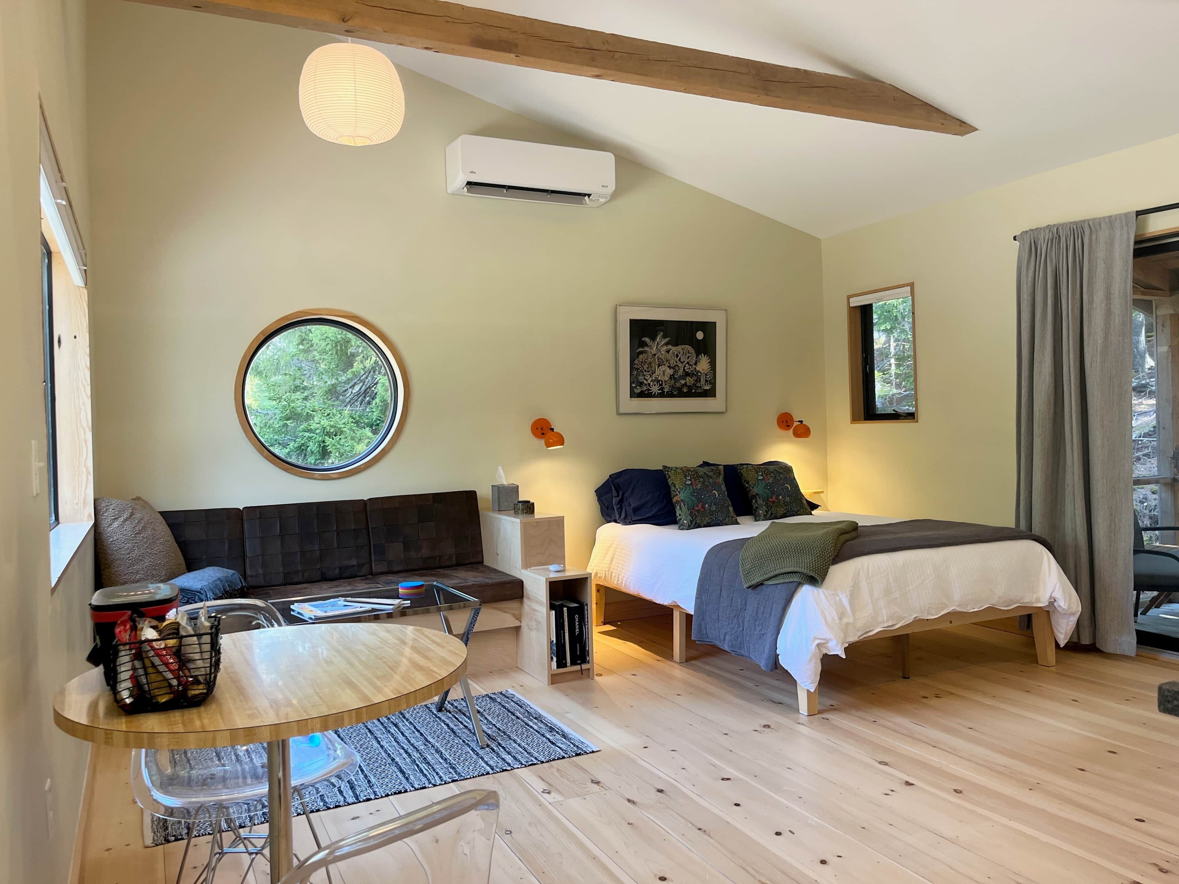 A room with a bed, a round window, a sofa, a table, and an air conditioning unit.