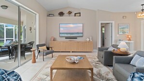 A modern living room with a large flat-screen TV, wooden furniture, and a glass door leading to an outdoor area. - Bonitaville | Coastal Family Oasis w/ Heated Pool & Outdoor Living (Bonita Springs)