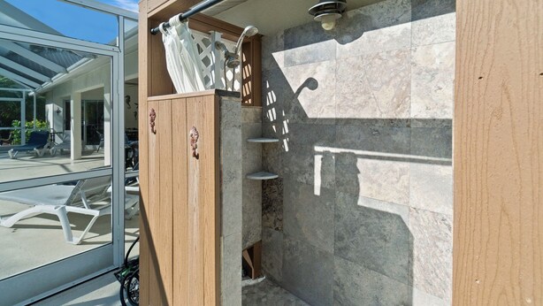 A wooden shower enclosure with a glass door, a showerhead, and a wall-mounted light fixture. - Bonitaville | Coastal Family Oasis w/ Heated Pool & Outdoor Living (Bonita Springs)