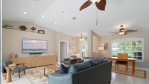 A spacious living room with a large gray sofa, a wooden coffee table, and a television mounted on the wall. There are ceiling fans, recessed lighting, and a view of the outdoors through large windows. - Bonitaville | Coastal Family Oasis w/ Heated Pool & Outdoor Living (Bonita Springs)