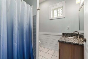 Une salle de bain avec un rideau de douche, une fenêtre, un lavabo doté d’un plan de travail en granit et un carrelage au sol.