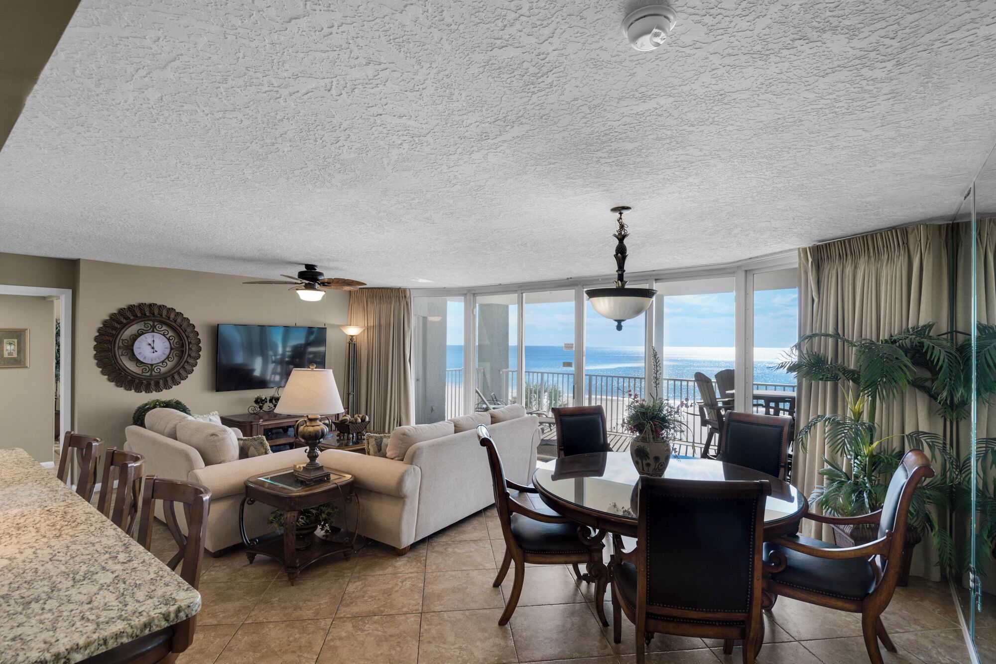 A spacious living room with a large window overlooking the ocean, a dining area with a round table, and a ceiling fan.