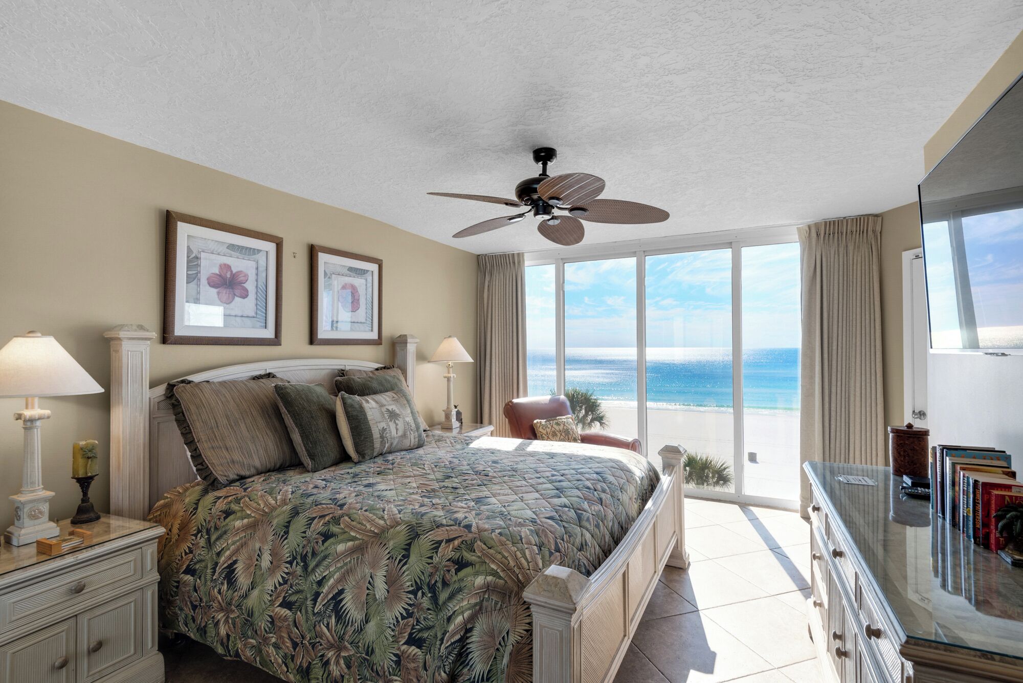 A bedroom with a large bed, a ceiling fan, a view of the ocean, and a balcony with books.