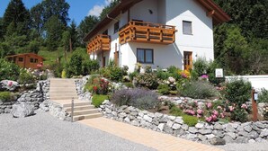 A two-story chalet with a balcony, surrounded by a garden with various plants and flowers. - Apartment 'Am Rosengarten' with Wi-Fi and Air Conditioning (Füssen)