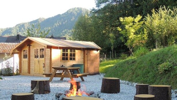 A wooden cabin with a fire pit surrounded by tree stumps, set on a gravel surface with a picnic table nearby. - Apartment 'Am Rosengarten' with Wi-Fi and Air Conditioning (Füssen)