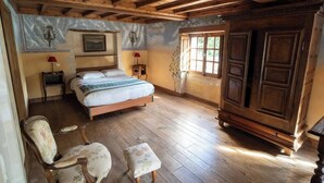 Een slaapkamer met een houten bed, een nachtkastje, een stoel, een klein krukje, een garderobe en een raam met uitzicht op groen.