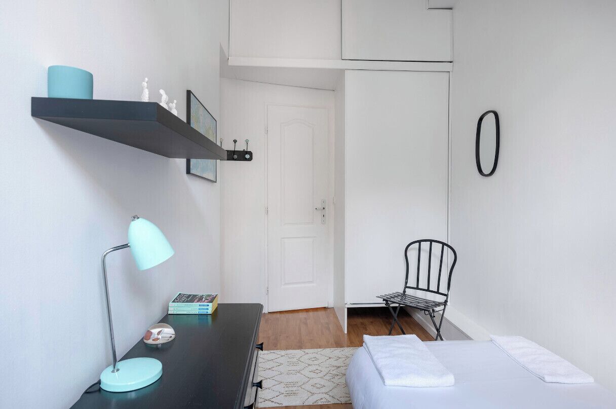 A small, neatly organized room with a bed, a desk with a lamp, a chair, and a shelf with decorative items.