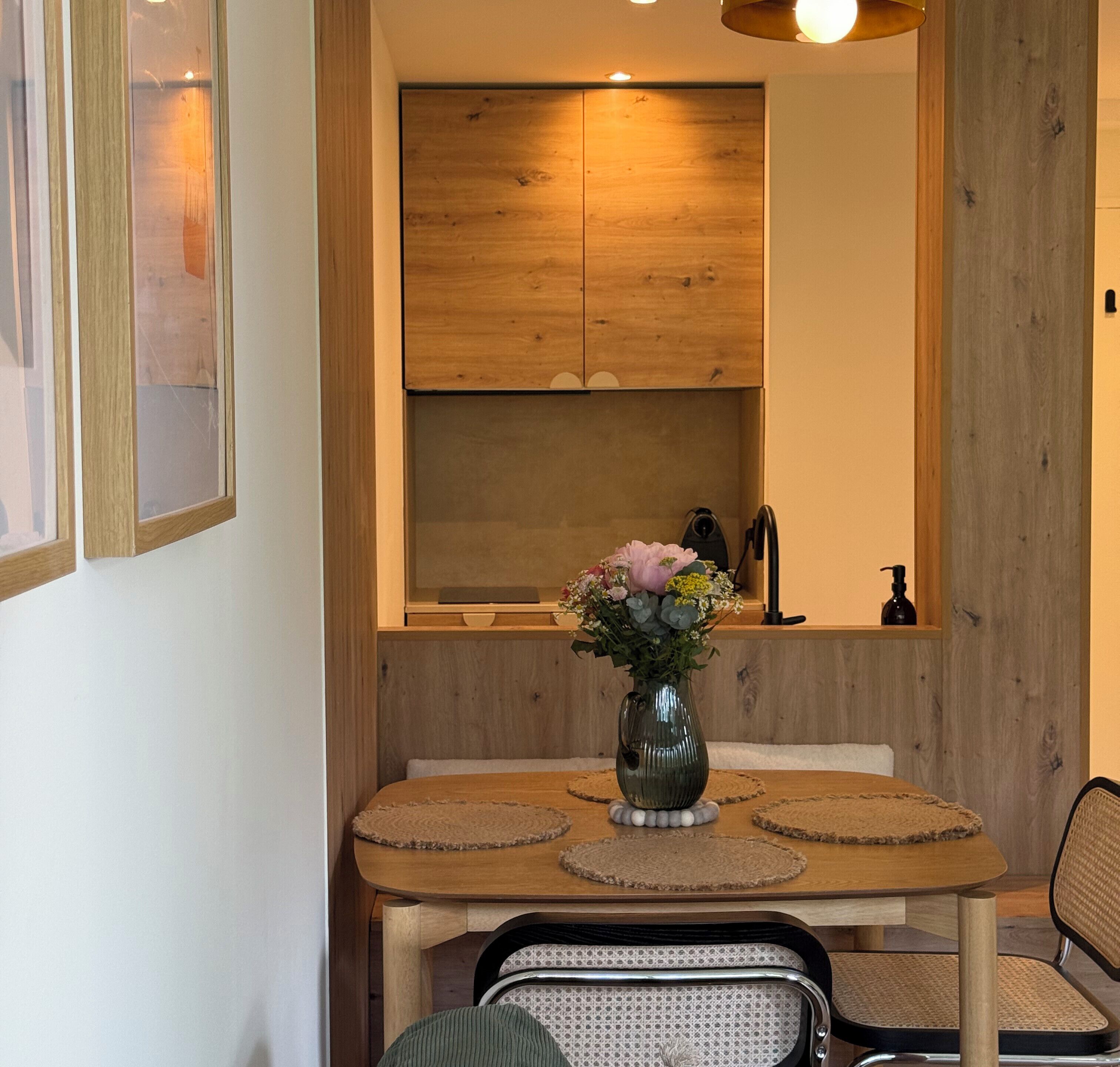Zona pranzo con tavolo in legno, sedie, un vaso con fiori e un mobiletto in legno sopra.