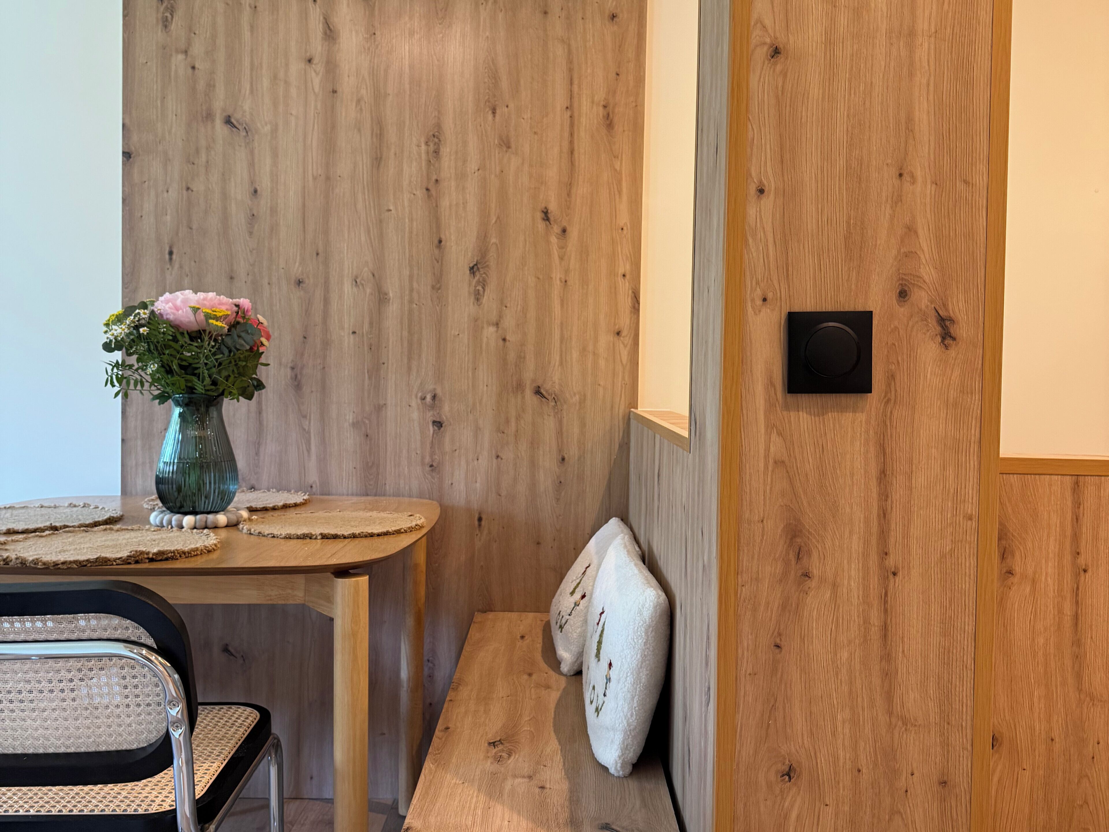 Una pared de madera, una pequeña mesa redonda con un florero, un banco y una silla.