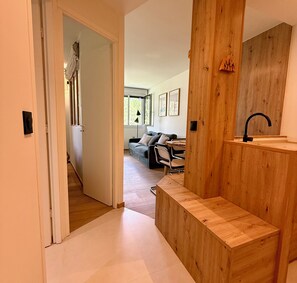 A modern interior with a wooden bench, a sink, and a view into a living area with a sofa and chairs. - Cosy apartment in the heart of Chamonix (Chamonix-Mont-Blanc)