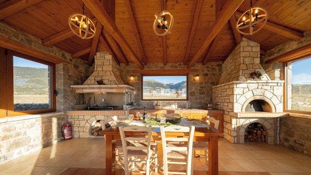 A rustic stone kitchen with a stone fireplace, wooden ceiling beams, and a large window overlooking a mountainous landscape. - Anemos Mountain Villa - Your Perfect Mountain Retreat (Arachova)