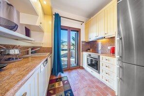 Una cocina con un refrigerador de acero inoxidable, una estufa y un lavamanos. Hay una ventana con cortina y una puerta que da al exterior.