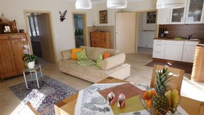 A living room with a sofa, a dining table with a fruit bowl, and a kitchen area with cabinets.