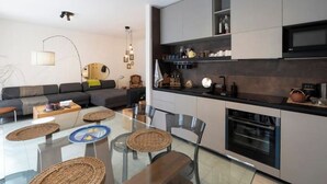 Eine moderne Küche mit Einbauofen, ein Essbereich mit Glastisch und Stühlen sowie ein Wohnzimmer mit Sofa und Lampe.