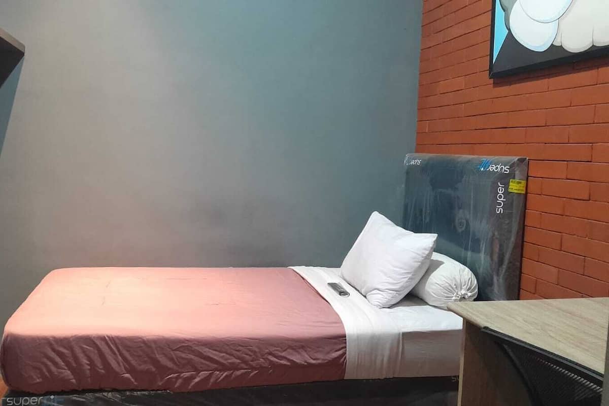 A single bed with a pink bedspread and white pillows, a small desk with a computer, and a wall-mounted television.