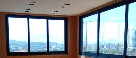 A spacious room with multiple wooden tables and chairs, large windows offering a city view, and recessed ceiling lighting.