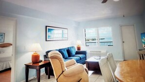 A living room with a blue sofa, a wooden coffee table, and a wicker chair. There is a lamp on a side table, and a framed picture on the wall. - Key Largo Waterfront 1 bedroom condo. (Key Largo)