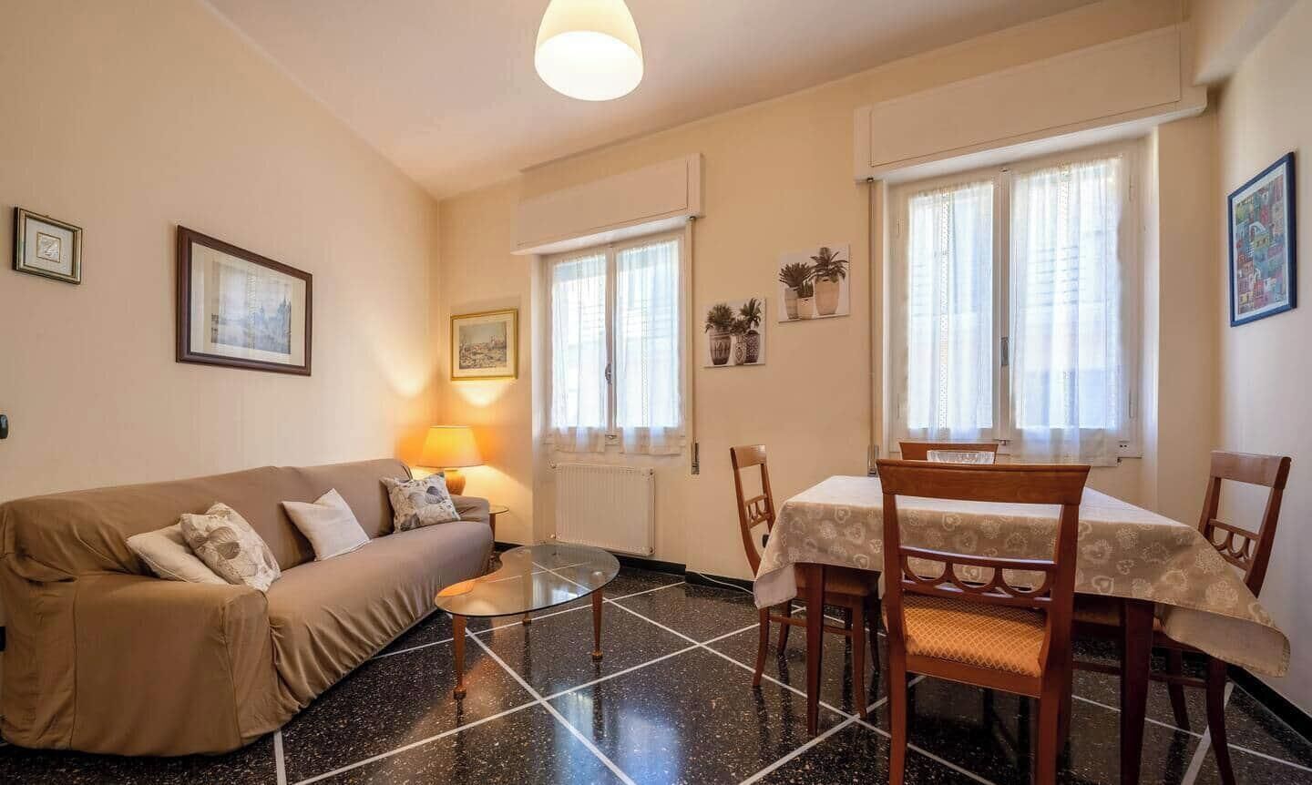 Un soggiorno con un divano, un tavolino in vetro e un tavolo da pranzo con sedie. Sono presenti due finestre con tende, un termosifone e quadri appesi alle pareti.