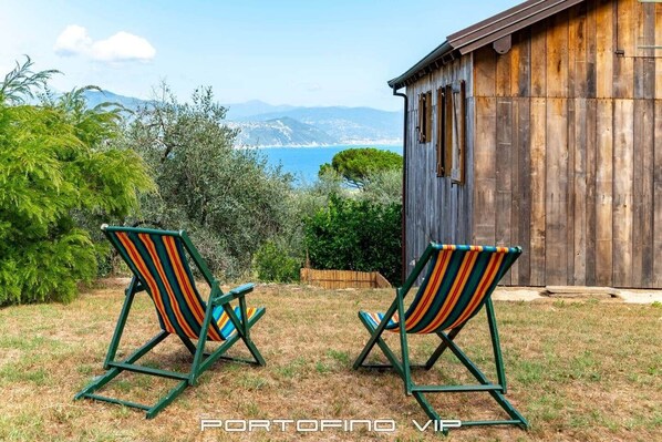 Zwei gestreifte Liegestühle auf einer Rasenfläche mit einem Holzgebäude im Hintergrund und Blick auf das Meer und die Berge.
