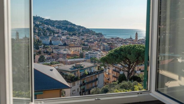 A coastal cityscape viewed from a window, featuring a mix of residential and commercial buildings, a prominent hill with a tower, and a clear view of the sea. - Cosy Apartment by PortofinoVip [Parking-Seaview] (Rapallo)