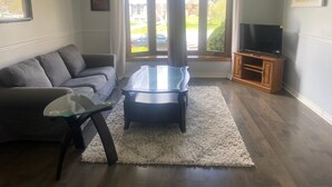 A living room with a sofa, glass coffee table, TV stand, and large windows. - Home Away from Home - Relax and unwind in this beautifully decorated 3-bedroom (Kingston)