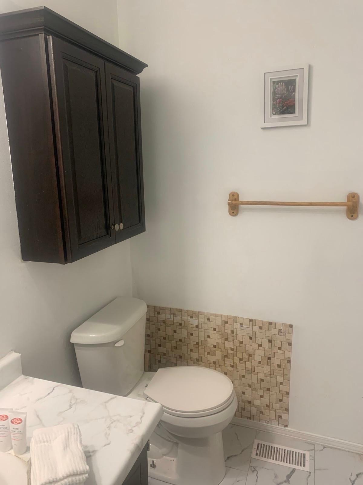 A bathroom with a toilet, a vanity with a marble countertop, a dark wooden cabinet, a small framed picture on the wall, and a beige mosaic tile backsplash.