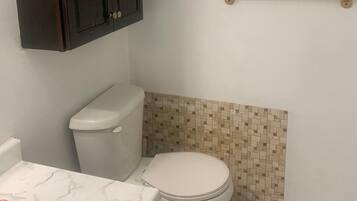 A bathroom with a toilet, a vanity with a marble countertop, a dark wooden cabinet, a small framed picture on the wall, and a beige mosaic tile backsplash.