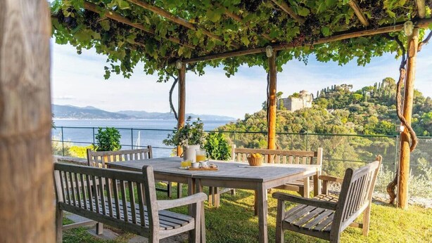 Ein Außenbereich mit Tisch und Stühlen, einer mit Wein bewachsenen Pergola sowie Meerblick und einer hügeligen Landschaft im Hintergrund.