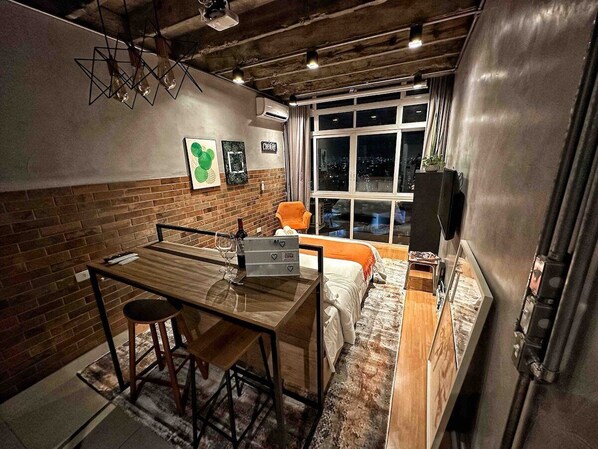 Une pièce de taille modeste, avec une table en bois, une seule chaise orange, un lit recouvert d’une couverture blanche et une fenêtre donnant sur la ville.