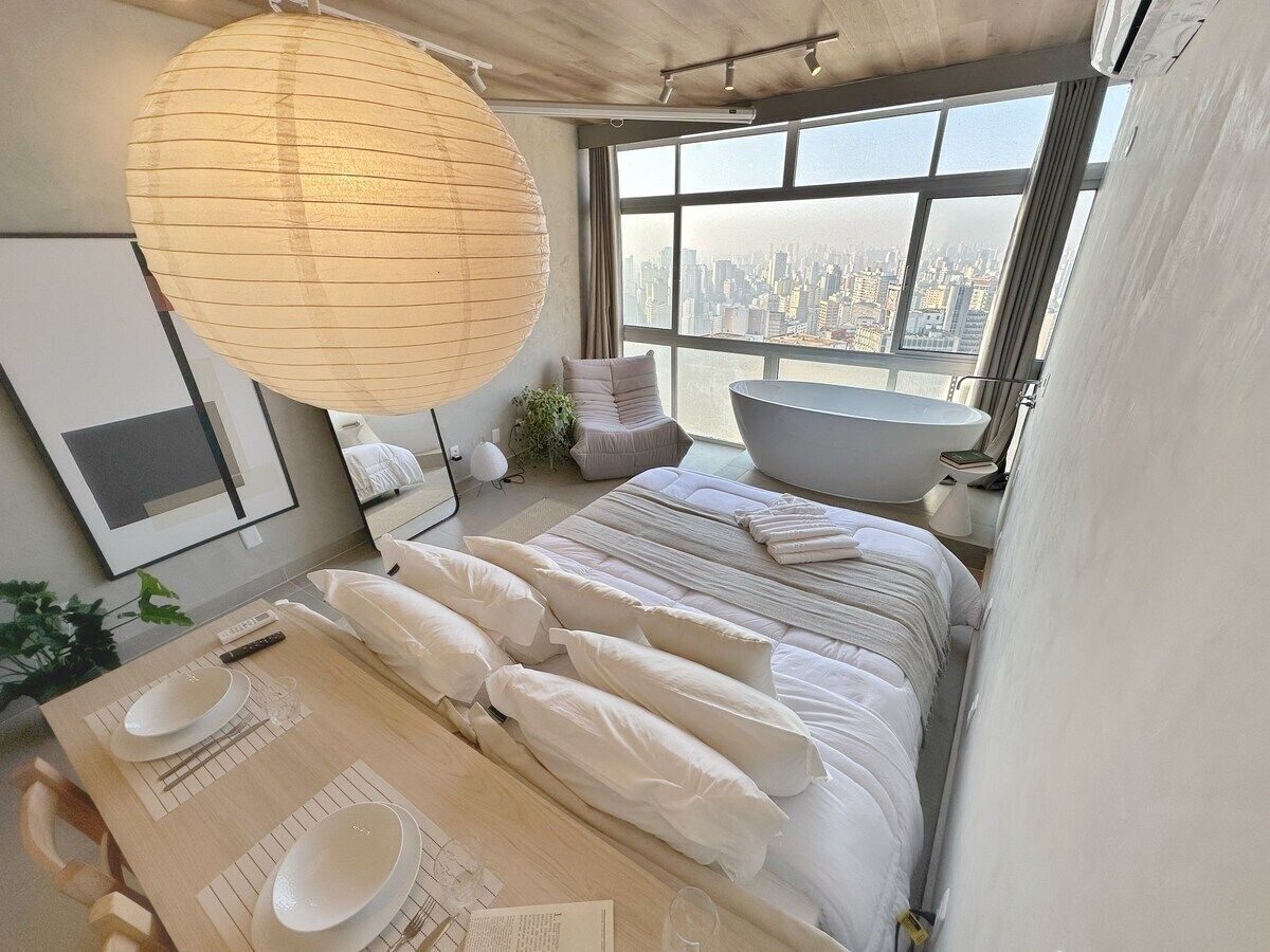 Une chambre moderne avec vue sur la ville, un grand lit avec une literie blanche, une table de chevet avec une lampe et un luminaire suspendu.