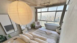 Et moderne soveværelse med udsigt over byen, en stor seng med hvide sengetæger, et natbord med en lampe og en hængende loftlampe.