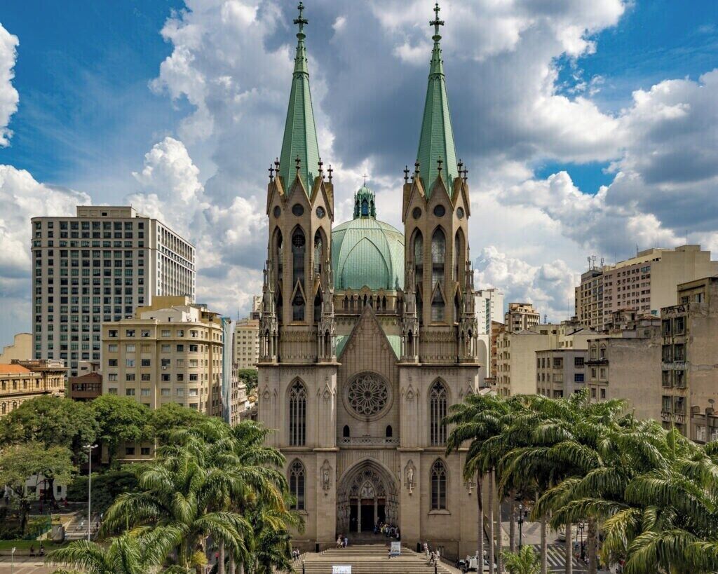 A cathedral with two tall spires and a large dome, surrounded by palm trees and modern buildings.