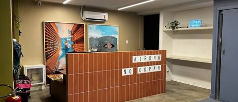 Un espace d’accueil avec un panneau de séparation marron sur lequel est inscrit « AIRBNB NO COPAN » et une peinture représentant un paysage urbain.