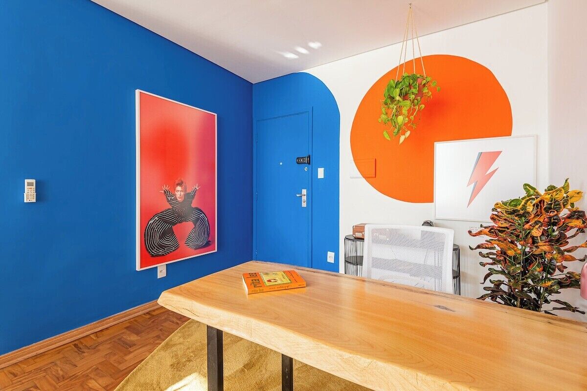 A room with a blue wall, a wooden table, a red artwork, a white wall with an orange circle, and a hanging plant.