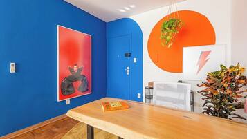 A room with a blue wall, a wooden table, a red artwork, a white wall with an orange circle, and a hanging plant.