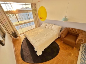 A bedroom with a bed, a brown leather armchair, a large window, and a hanging light fixture.