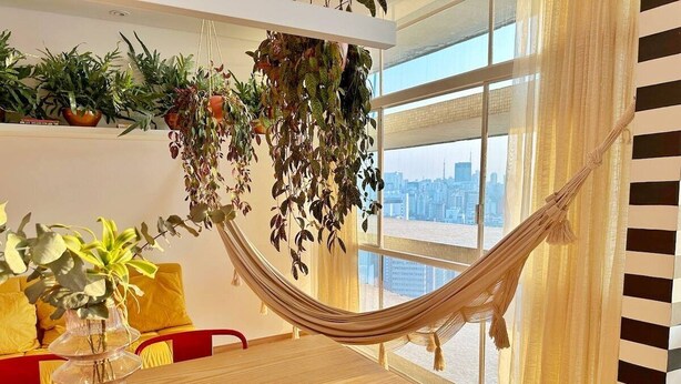 A room with a wooden table, a hammock, and a view of a cityscape through large windows.