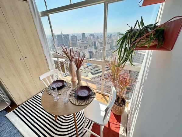 Un coin repas avec une table dressée pour deux personnes, une plante en pot et une vue sur le paysage urbain à travers de grandes fenêtres.
