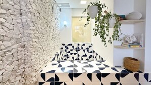 A bathroom with a stone wall, a geometric patterned floor, a hanging plant, and a shelf with books and a basket. - 1609B (São Paulo)