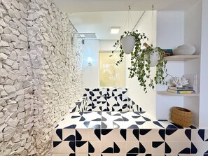 A bathroom with a stone wall, a geometric patterned floor, a hanging plant, and a shelf with books and a basket. - 1609B (São Paulo)