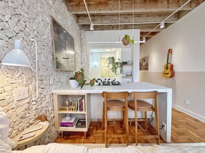 A cozy room with a stone wall, a white desk with two chairs, a guitar, and a hanging plant. - 1609B (São Paulo)