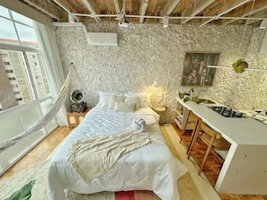 A bedroom with a large bed, a hammock, a small table with a lamp, and a kitchenette area. - 1609B (São Paulo)