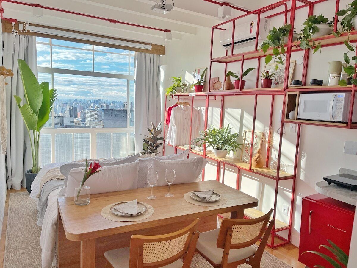 A dining area with a table set for two, a city view through a window, and a red shelving unit with plants.
