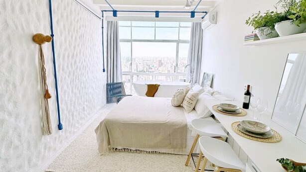 Un dormitorio con cama, escritorio, silla, ventana y planta en la pared, ofreciendo vistas a la ciudad.