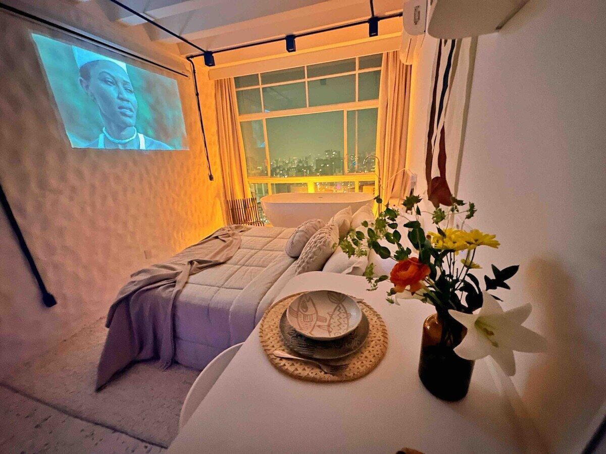 A room with a bed, a dining table with a vase of flowers, and a projection screen on the wall.