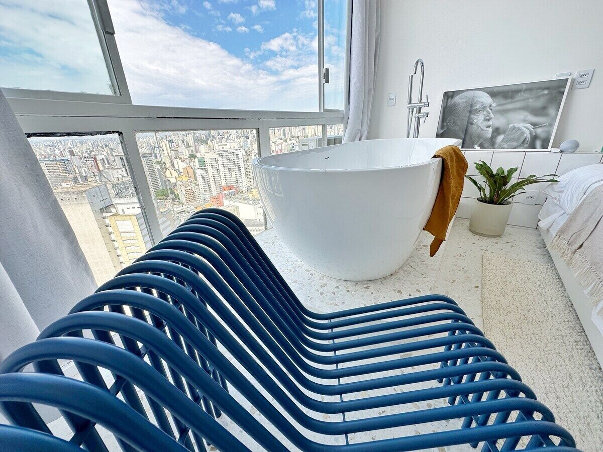 A modern bathroom with a large bathtub, a city view through a large window, and a blue chair.