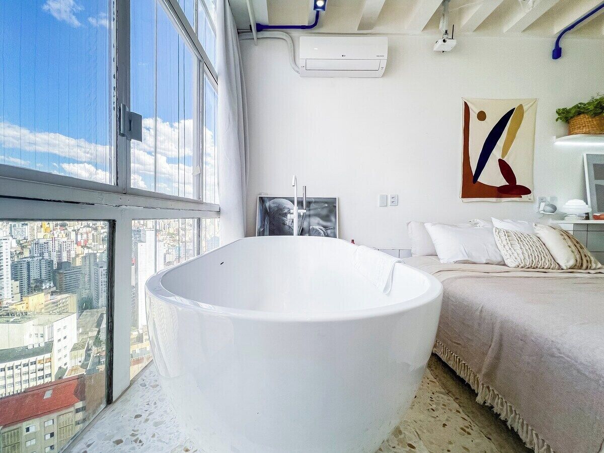 Ein modernes Badezimmer mit einer großen Badewanne, ein Bett mit einer beigefarbenen Tagesdecke und ein Blick auf die Stadt durch ein großes Fenster.
