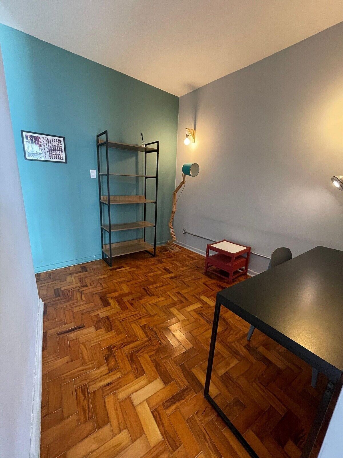 A room with a herringbone wooden floor, a metal shelving unit, a desk with a chair, and a wall-mounted lamp.