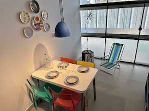A dining area with a table set for four, colorful chairs, a blue pendant light, and a view of the outside through large windows. - 0054E (São Paulo)