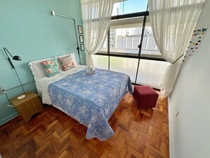 A bedroom with a bed, a nightstand, a window with curtains, and a view of buildings outside. - 0054E (São Paulo)